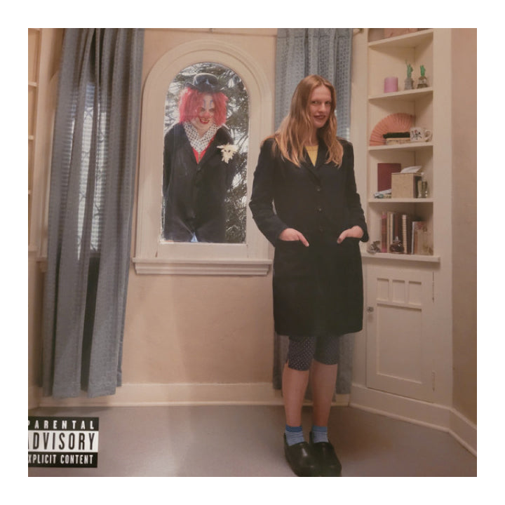 A woman stands in a room wearing a long black coat, patterned tights, blue socks, and black shoes. A large portrait of a clown is visible in an arched window behind her, and a parental advisory label appears in the bottom-left corner.