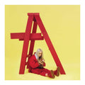 A person with platinum-blonde hair sits on the floor inside a large red wooden A-frame structure against a solid pale yellow background, wearing a matching red outfit and holding a gold chain.