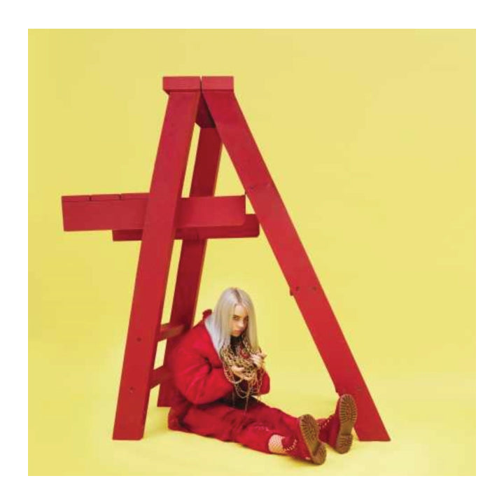 A person with platinum-blonde hair sits on the floor inside a large red wooden A-frame structure against a solid pale yellow background, wearing a matching red outfit and holding a gold chain.