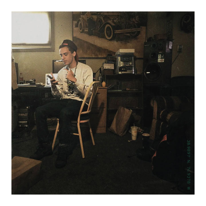 A man sits on a wooden chair in a dimly lit, cluttered room, holding a cigarette and looking down at a small object in his hand. The room contains shelves with equipment, a poster of a vintage car, and various other items.