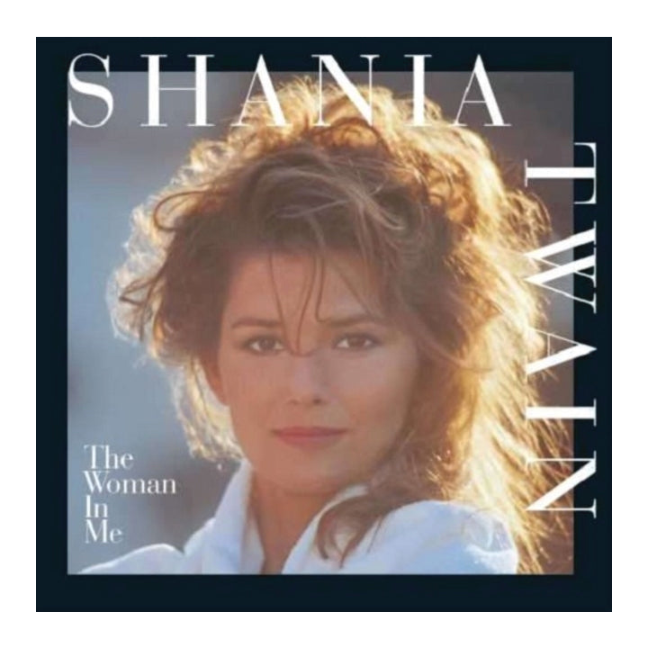 Album cover for Shania Twain's 'The Woman In Me', featuring a portrait of the artist with voluminous light-brown hair, wearing a white collared shirt. The text 'SHANIA TWAIN' is displayed prominently in white, and the album title is in the lower-left corner.