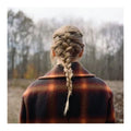 A person viewed from behind wearing a thick, multi-colored plaid coat with brown, orange, and black tones, featuring a detailed blonde braid hairstyle, set against a blurred outdoor background of bare trees.